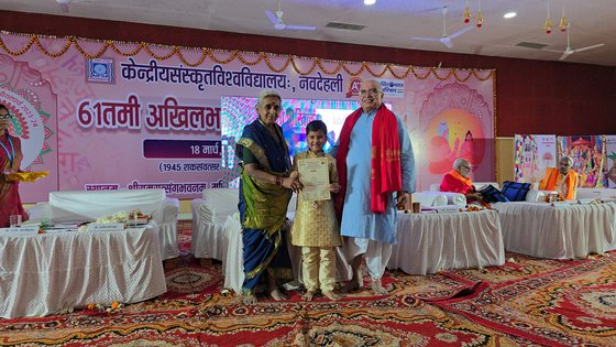 Vrishvardhan Sharma at six, holding his certificate between two elders on stage, under a banner reading Kendriya Samskrita Vishvavidyalayah, Navadehali, 61st Akhil Bharatiya competition.