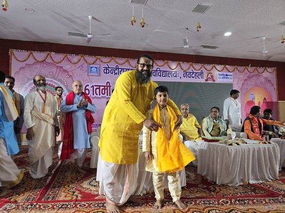 A yellow angavastram being placed around Vrishvardhan Sharma's shoulders on stage at the same competition, with the assembly applauding.