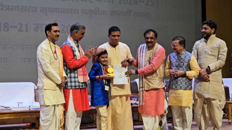 Vrishvardhan Sharma receiving his certificate on stage at the 62nd Akhil Bharatiya Shastrotsav, Patanjali University, Haridwar, with a hand resting on his head in blessing.