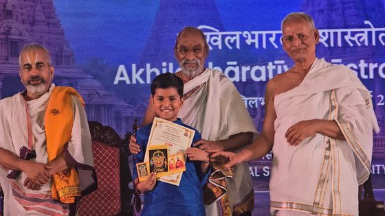 Vrishvardhan Sharma holding his first place certificate at the 63rd Akhil Bharatiya Shastrotsav, flanked by three scholars.