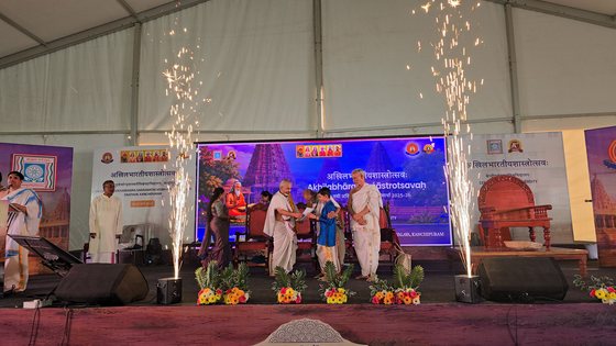 The award being handed over on stage between two fountains of sparks, under a banner reading Akhilabharatiyasastrotsavah, Sri Chandrasekharendra Saraswathi Viswa Mahavidyalaya, Enathur, Kanchipuram.