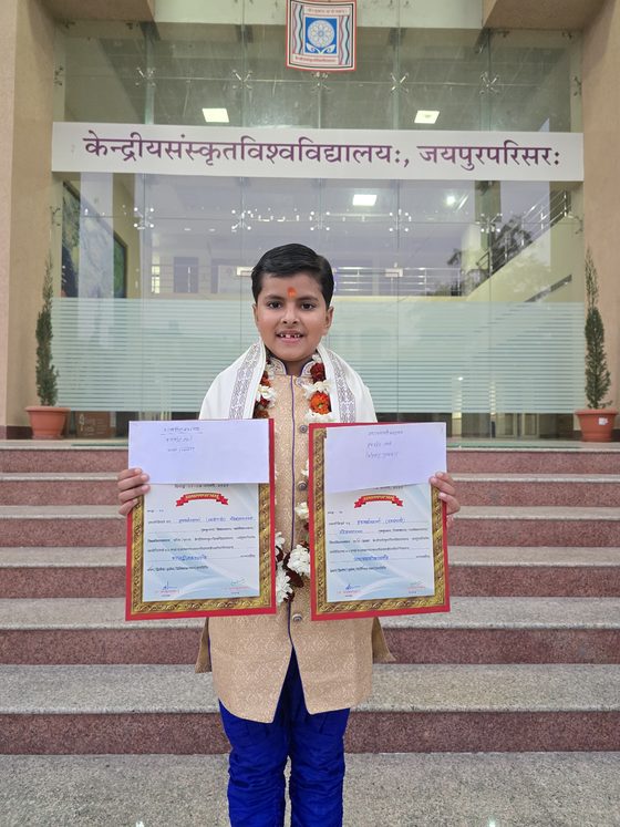 Vrishvardhan Sharma holding two certificates on the steps of the Central Sanskrit University Jaipur campus, January 2025.