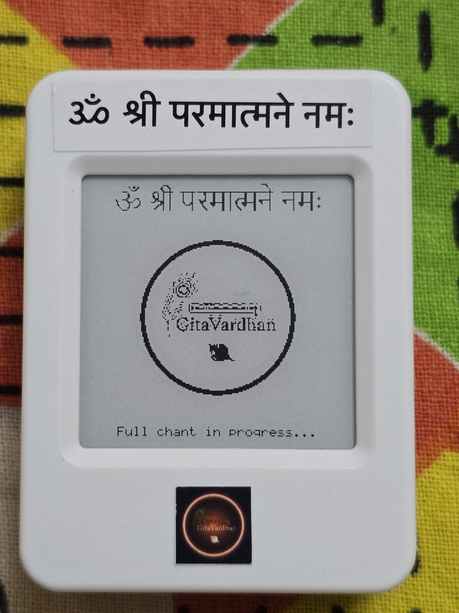 The e-ink device resting on printed cloth, its screen reading Om Sri Paramatmane Namah with the note, full chant in progress.