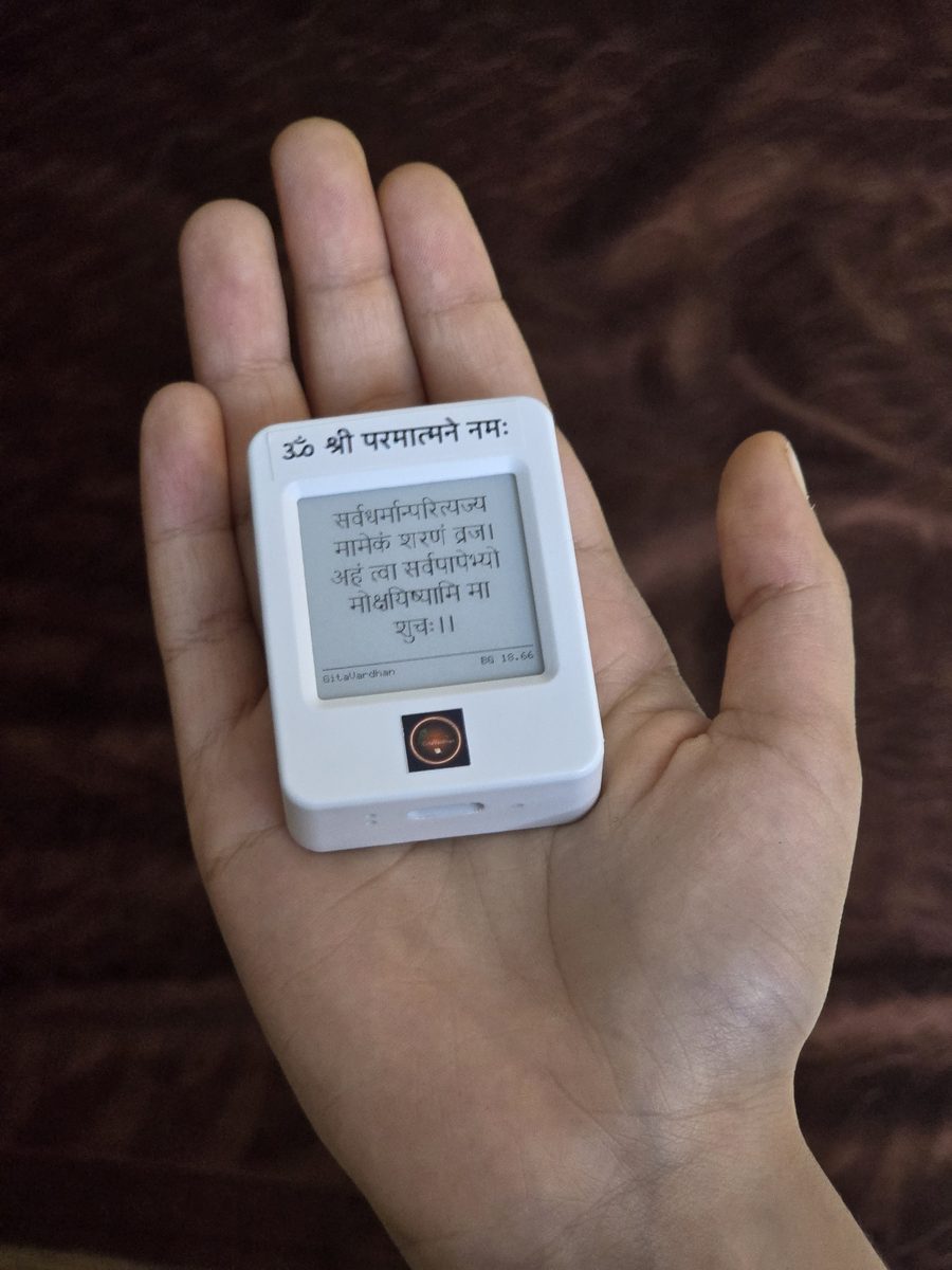 The GitaVardhan device held in an open palm, its e-ink screen showing Bhagavad Gita 18.66 in Devanagari.