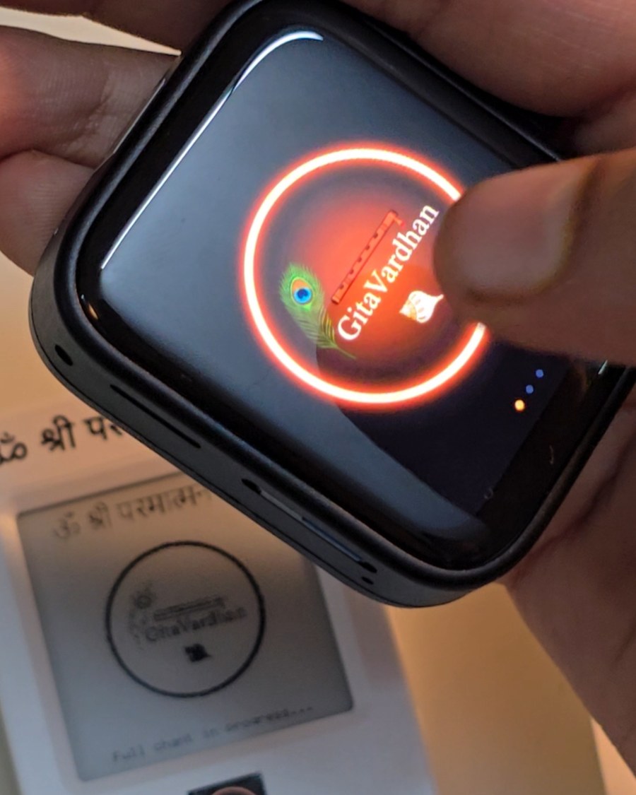 The touchscreen GitaVardhan device held close, its screen showing the GitaVardhan mark mid load, with the e-ink device visible on the table below it.