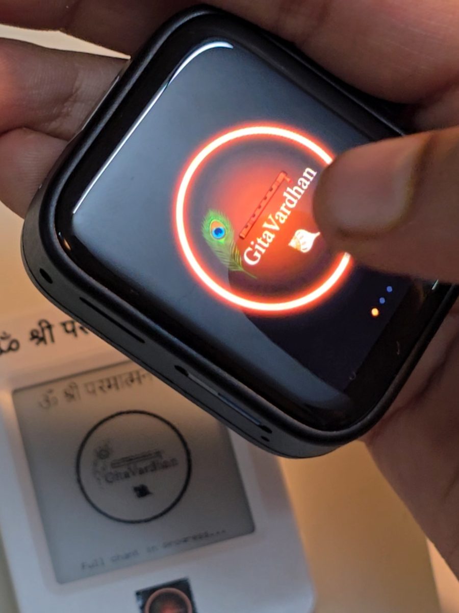 A phone running the GitaVardhan app held above the e-ink device, both showing the same mark.