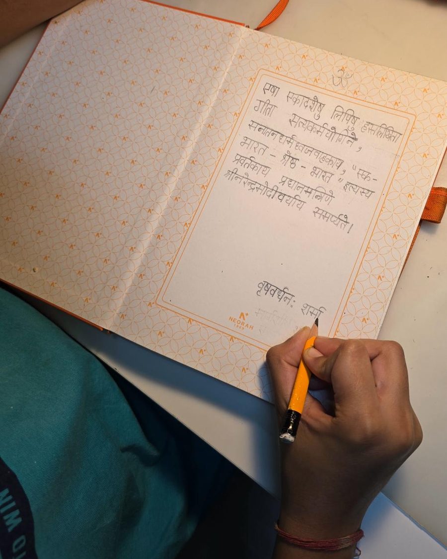 A hand writing a Sanskrit dedication in pencil on the opening page of a bound notebook.