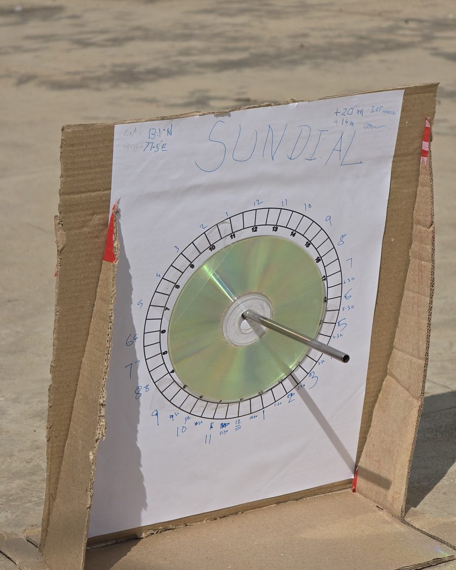 A handmade sundial: a compact disc and metal tube gnomon mounted on paper over cardboard, marked with latitude, longitude and time corrections.