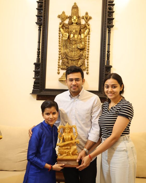 Vrishvardhan Sharma presenting a carved wooden Saraswati to Shri Tejasvi Surya, with Sushri Sivasri Skandaprasad beside them, beneath a brass relief.