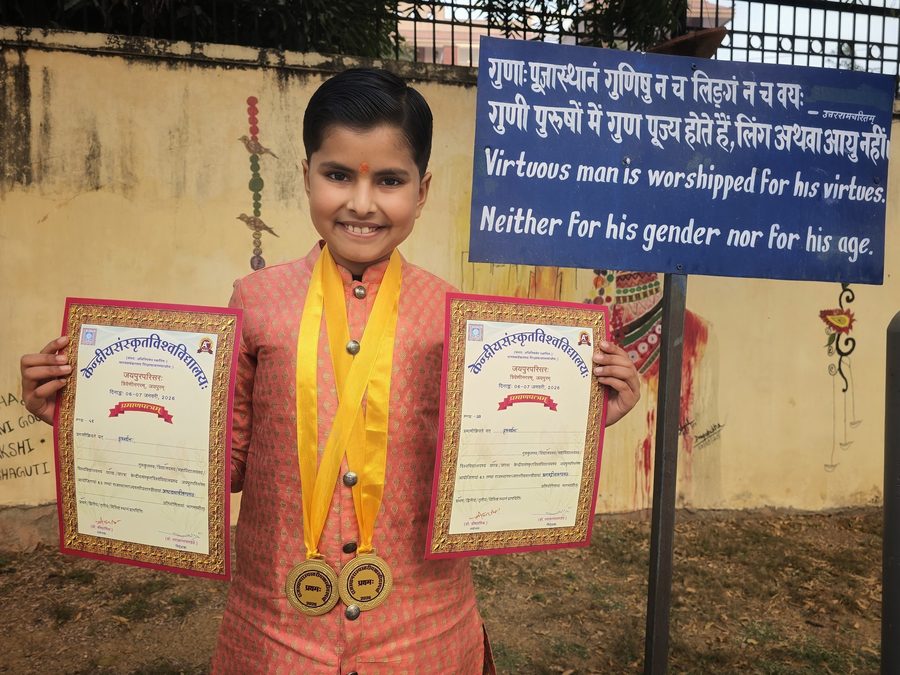 Vrishvardhan Sharma holding two certificates outside a competition venue, beneath a board bearing a Sanskrit verse and its English translation.