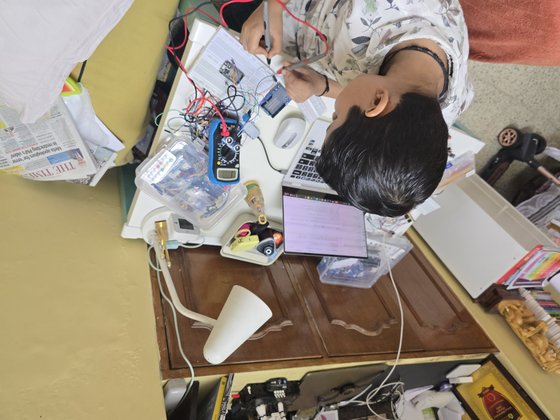 Vrishvardhan Sharma testing a circuit with a multimeter at his desk, laptop open beside a breadboard.