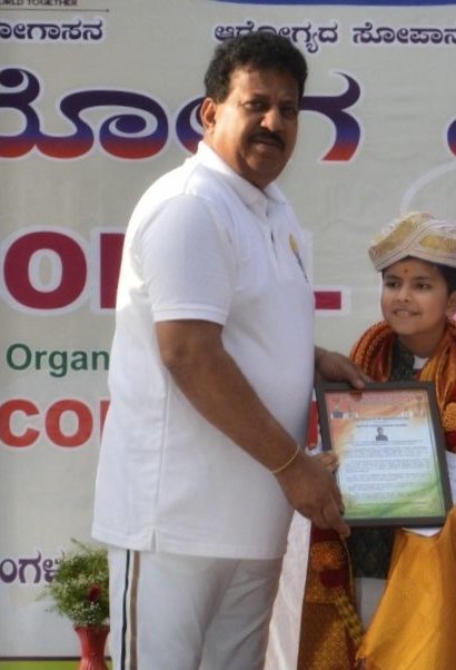 Shri S. R. Vishwanath, MLA, handing Vrishvardhan Sharma a framed Certificate of Appreciation on stage at the International Yoga Day event in Bengaluru, June 2026.